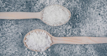 Some sea salt in wooden spoons on gray textured background, flat lay.